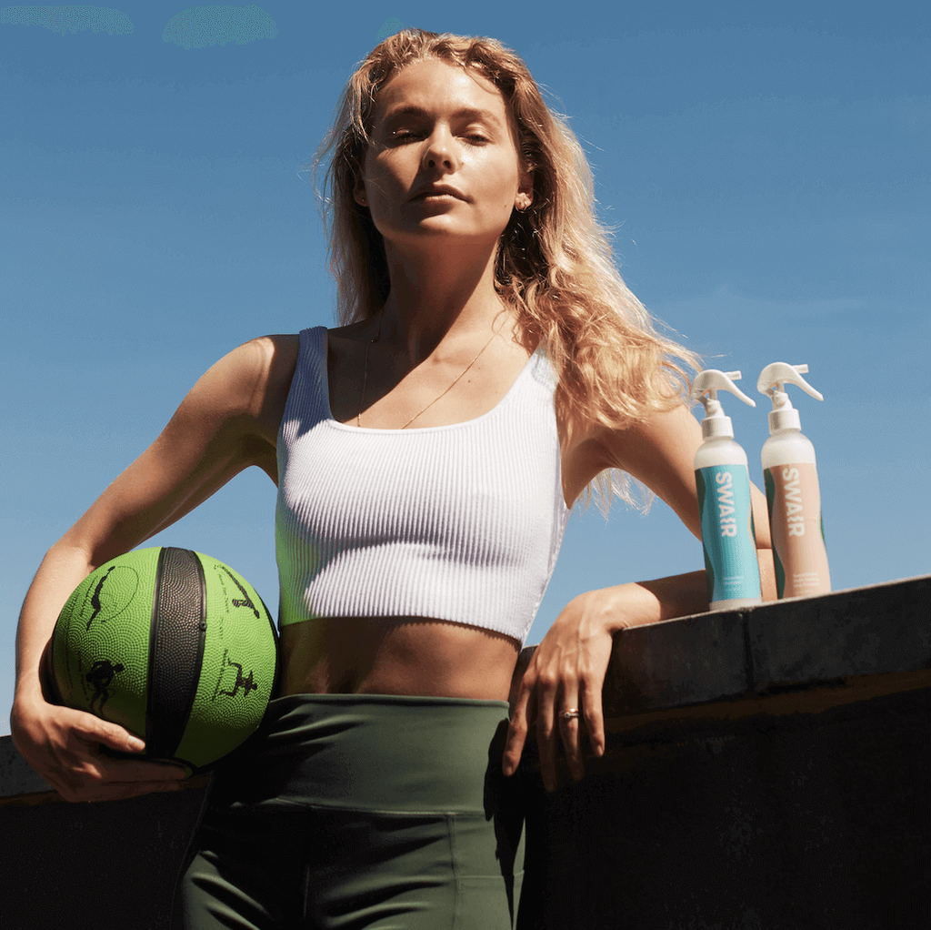 Model holding a medicine ball with Showerless Shampoo bottle and SweatShield Leave-In Conditioner bottle next to her
